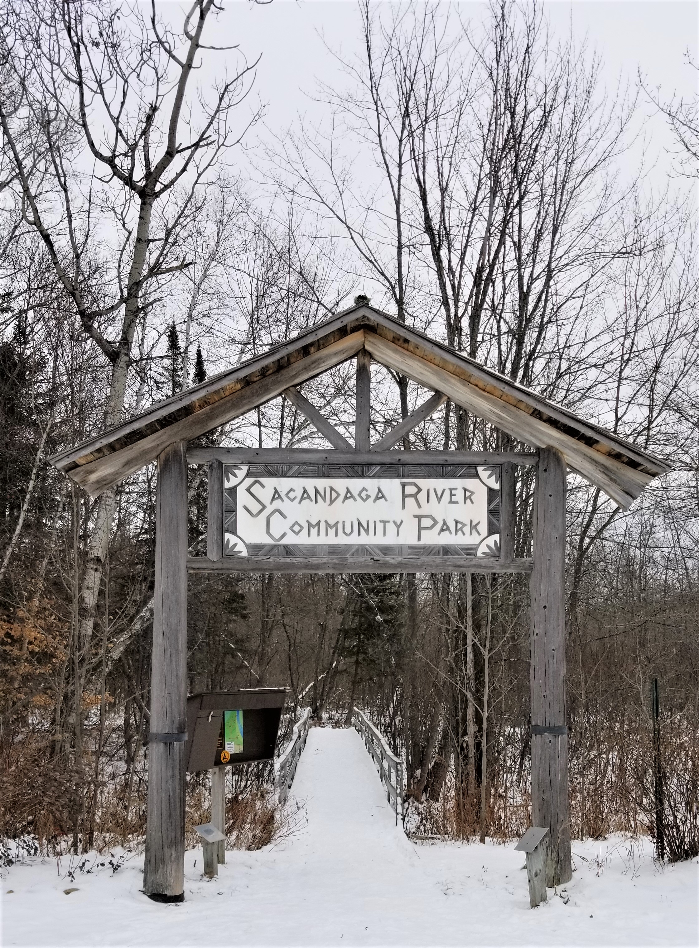 The entrance to the Sacandaga River Pathway. A large wooden frame with a pointed roof rises above the trail entrance. The sign on the frame reads, "Sacandaga River Community Park." The photos is taken in winter and the trees are bare of leaves and the ground is covered with snow.