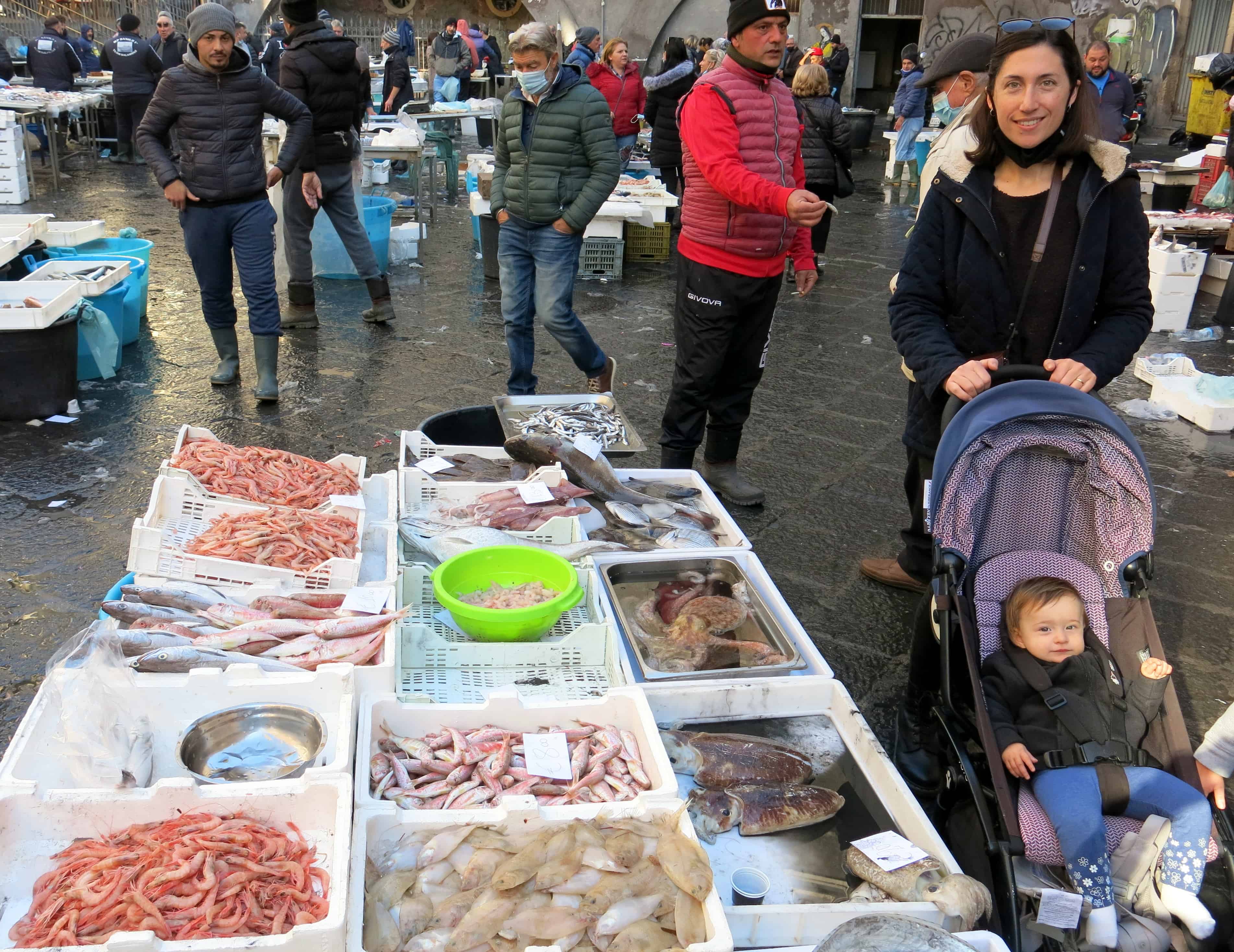 A photo of a mother and baby at the Catania fish market, standing next to tables full of fresh caught fish. Sicily.