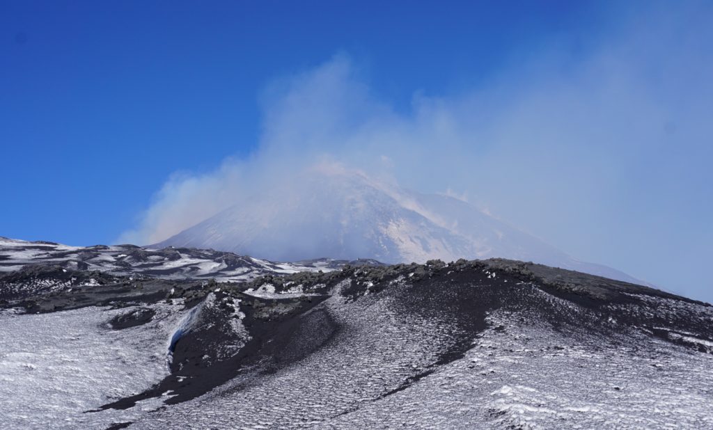 The summit of Mount Etna with a plume of smoke rising, set against a clear blue sky with patches of snow on the black volcanic ground, highlighting the contrast of nature's elements.