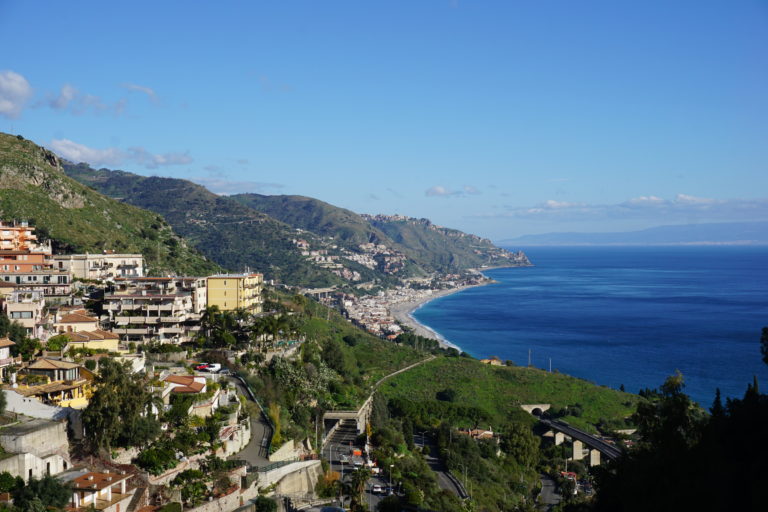 buildings on a hillside, with green rolling hills and the blue Mediterranean sea to the right.