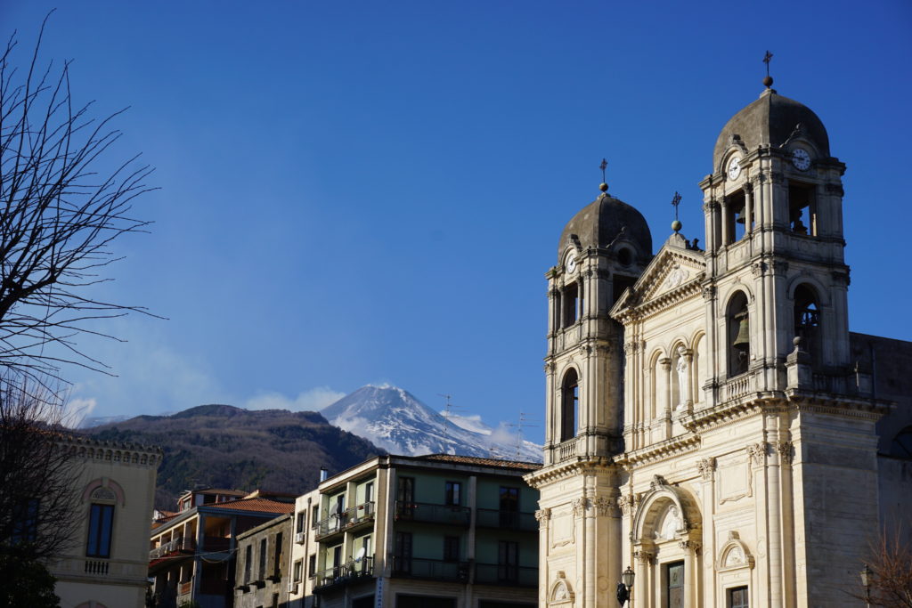 church with two bell towers and the snow covered and smoking peak of Mount Etna in the background.