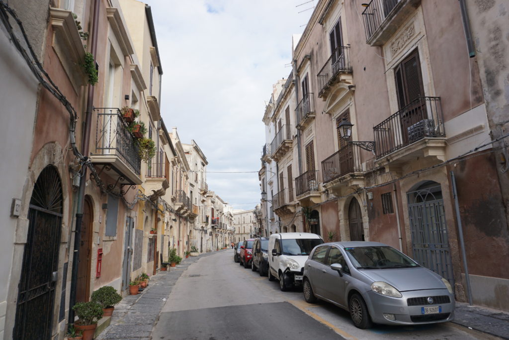 a narrow street lined on one side with parked cars. Pink buildings with black wrought iron balconies line either side.