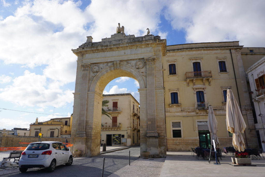 a white car driving through a stone gate in the city of Noto in Sicily.