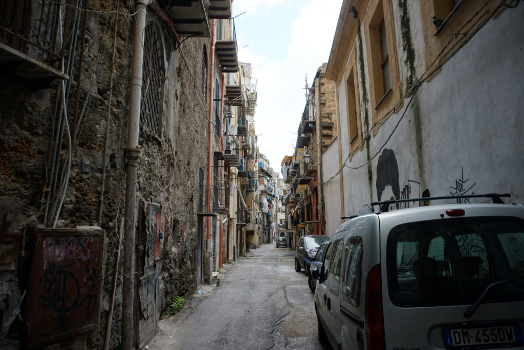 narrow alleyway with cars parked along one side. The buildings on either side tower over the street, blocking out most of the sunlight.