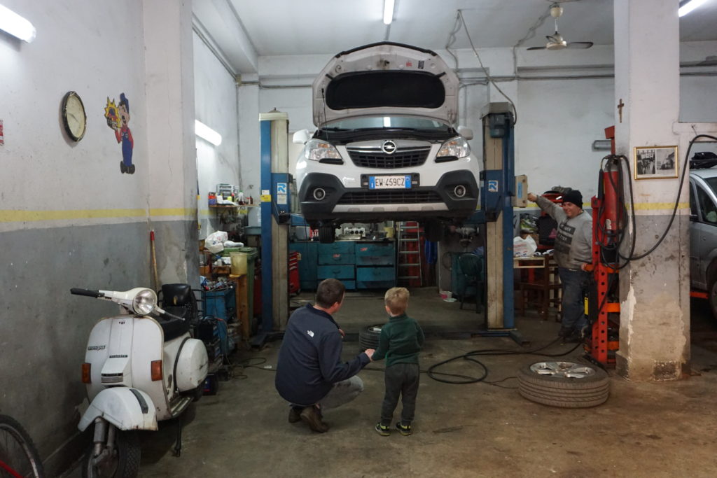 a man and young boy look up at a white car being lifted up in a mechanic's shop.