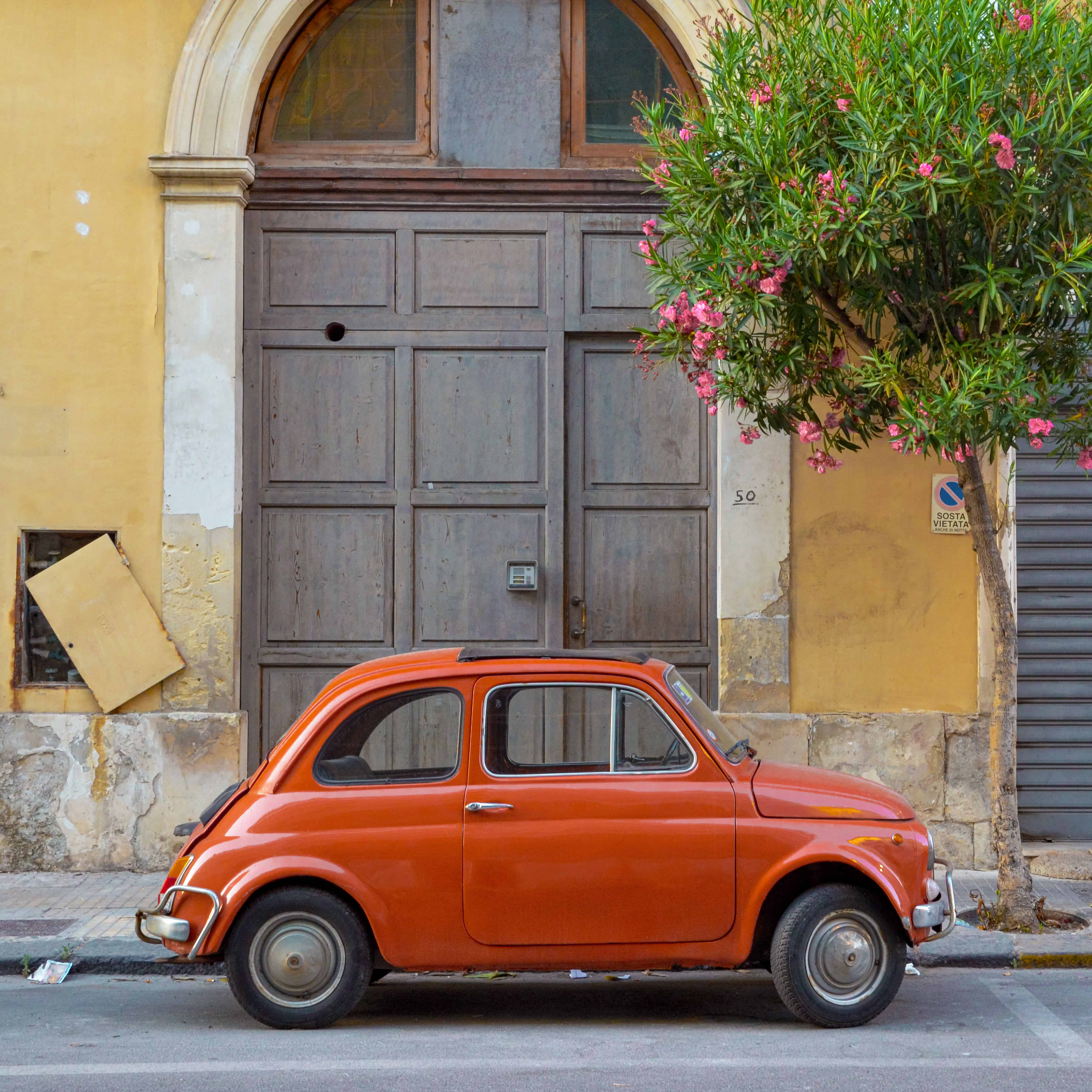 Photo of a cute, small red car in front of a wooden doorway and yellow wall.