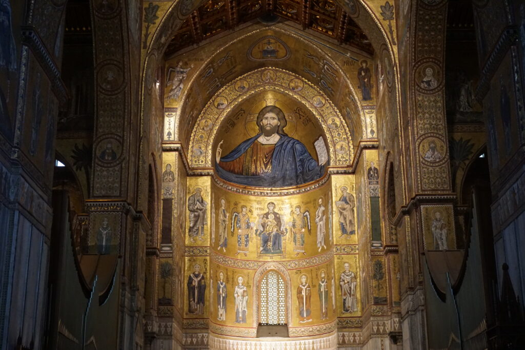 The interior of Monreale Cathedral in Sicily features towering Byzantine mosaics glowing with gold tiles that cover the apse and surrounding arches. At the center is a large image of Christ Pantocrator with a raised hand and open book, surrounded by saints angels and biblical figures arranged in vertical panels that emphasize the height and grandeur of the space.