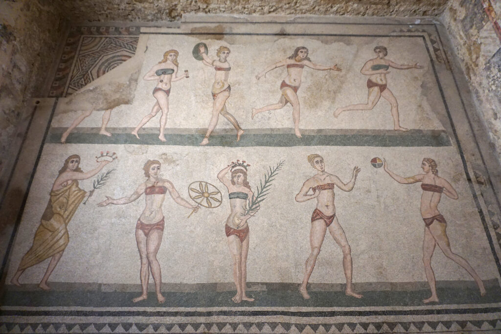 Ancient Roman mosaic showing women in two rows wearing bikini style garments while running jumping and holding sports equipment. The figures appear mid motion with items like a discus ball palm branches and crowns suggesting athletic competition. The mosaic is set into a stone floor with visible wear that highlights its age and historical context.