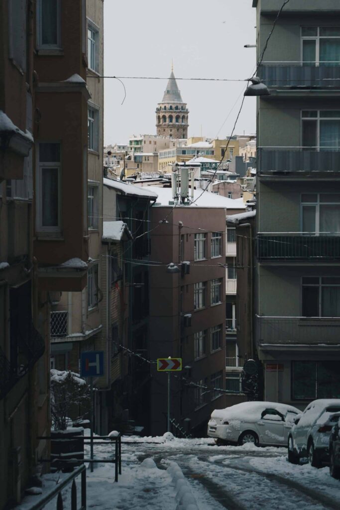 Istanbul's Galata Tower in the background, frames by snow covered building and cars in the foreground.