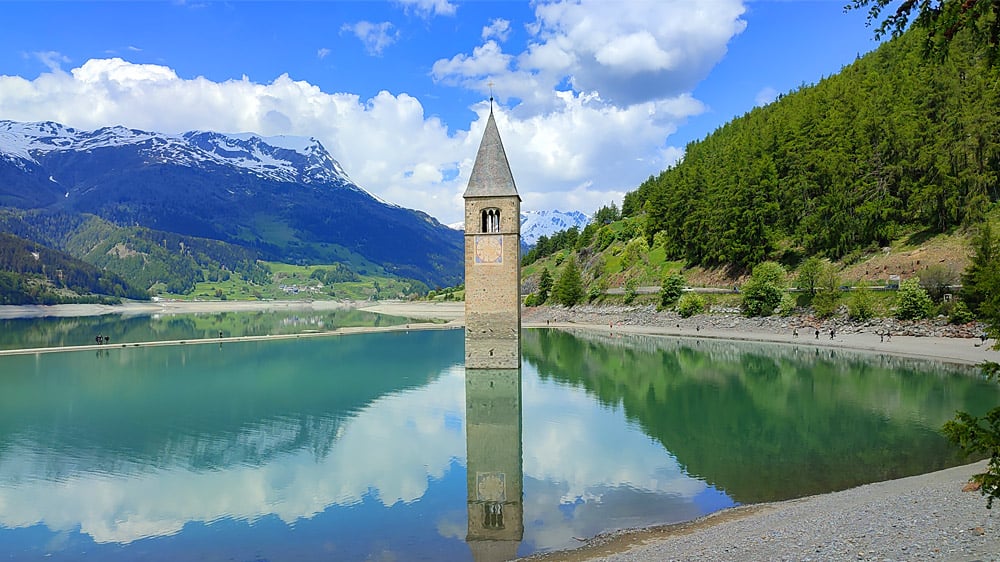 clock tower in the middle of a blue lake with snow-capped mountain peaks in the distance.