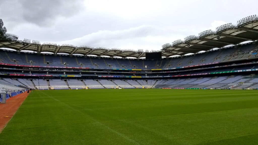 The expansive interior of Croke Park Stadium in Dublin, showcasing rows of empty seats in various hues under an intricate network of metal floodlights. The lush green pitch is impeccably maintained, ready for a Gaelic games match, set against an overcast sky.