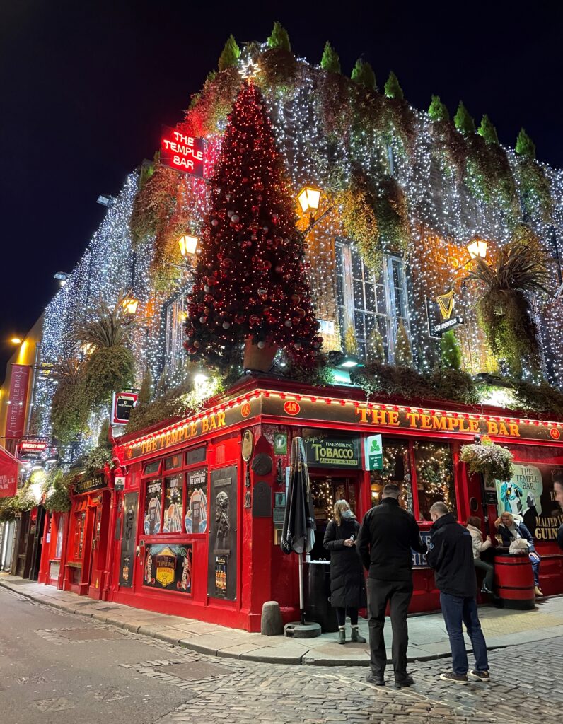 The Temple Bar pub in Dublin adorned with twinkling Christmas lights and decorations, creating a festive ambiance on a lively street at night. The iconic red exterior is complemented by a large Christmas tree, with people gathered outside, adding to the holiday cheer.