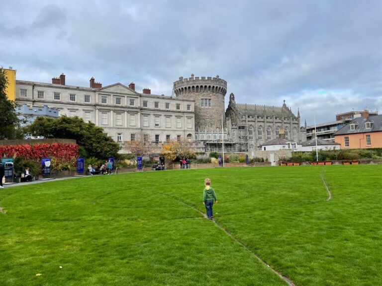 A child is seen walking across a lush green lawn towards Dublin Castle, a historic fortress with a prominent round tower and grey stonework. The castle stands under a cloudy sky, surrounded by well-maintained gardens and visitors enjoying the grounds.