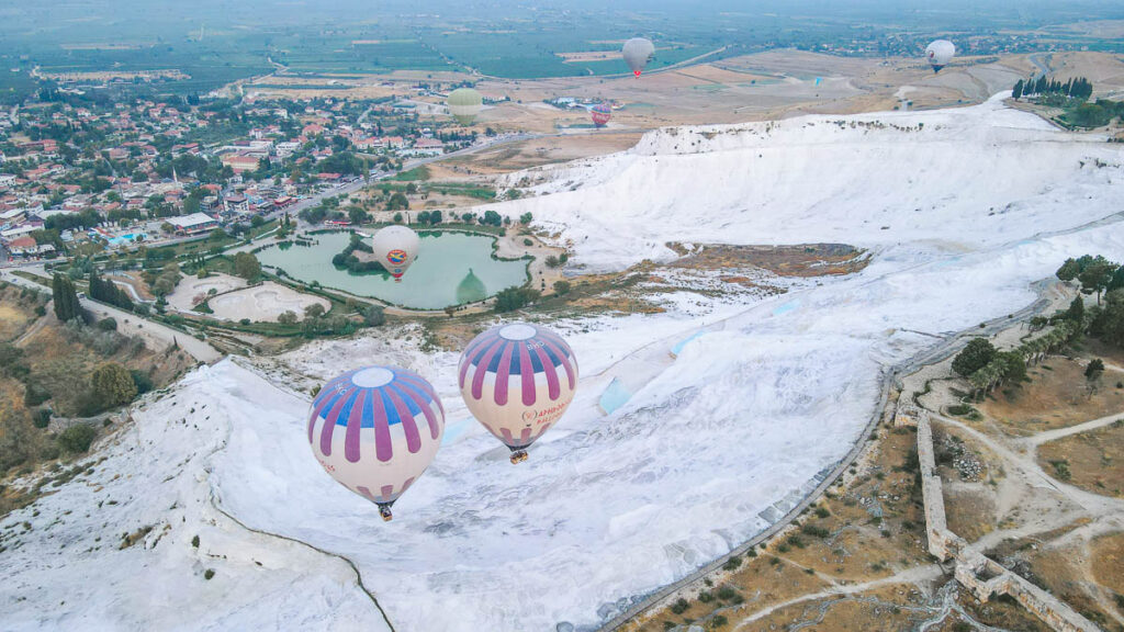 hot air balloons fly over white travertine pools of pamukkale.