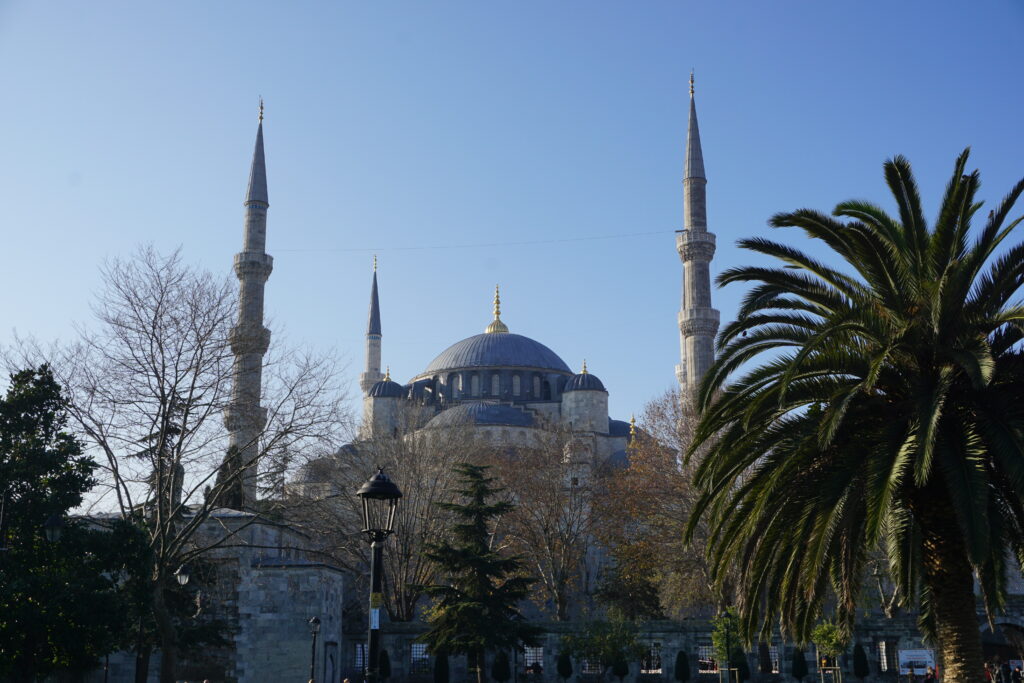 the blue mosque in istanbul, with a blue dome and three visible minarets, partially hidden behind trees.