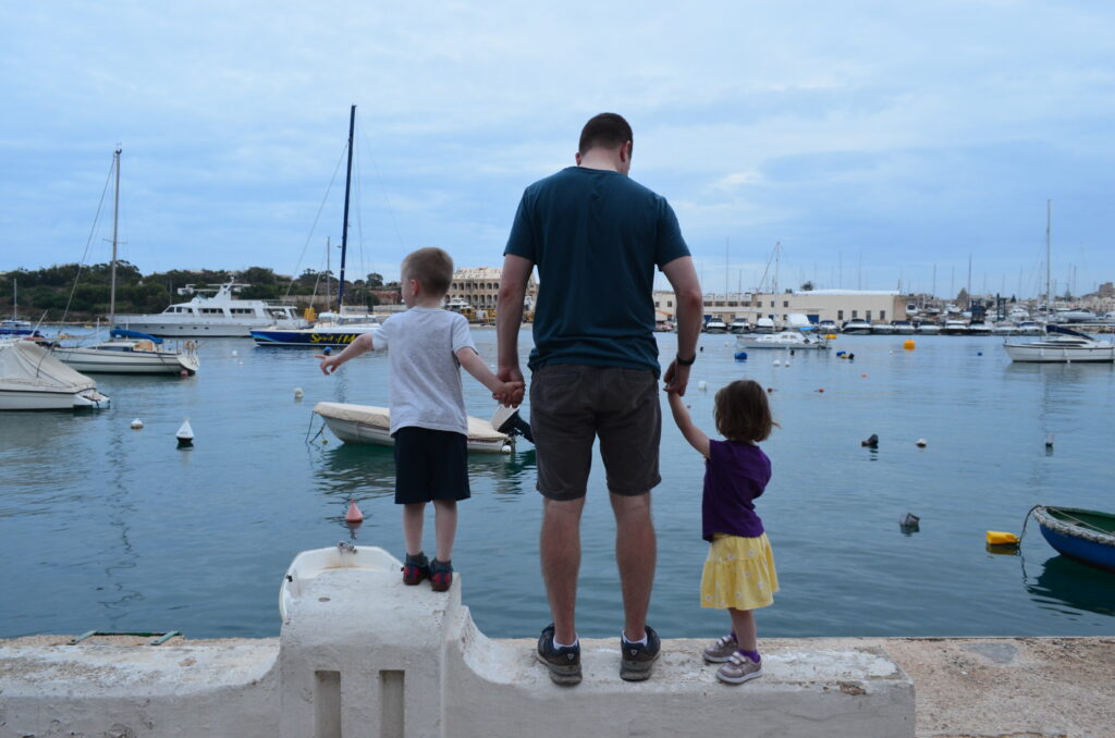 the author's husband stands on a wall in Malta with his kids. The man and his two children, one on either side of him, face away from the camera and look out on the harbor which is filled with boats.