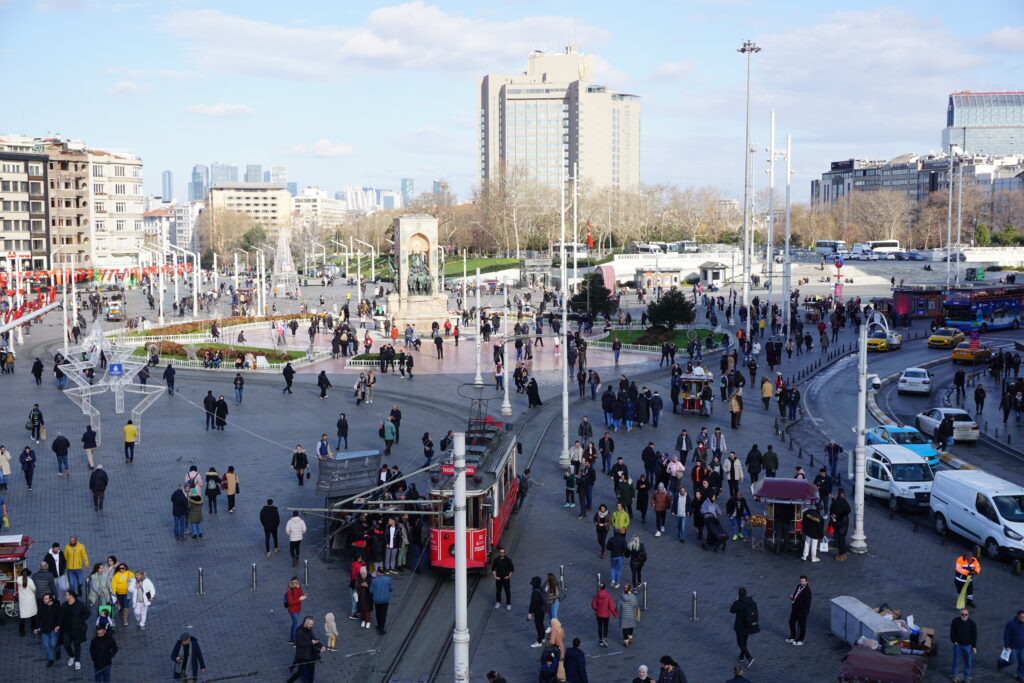 taksim square in istanbul, busy with a tram, people walking, a monument in the center, and large city buildings in the background. 