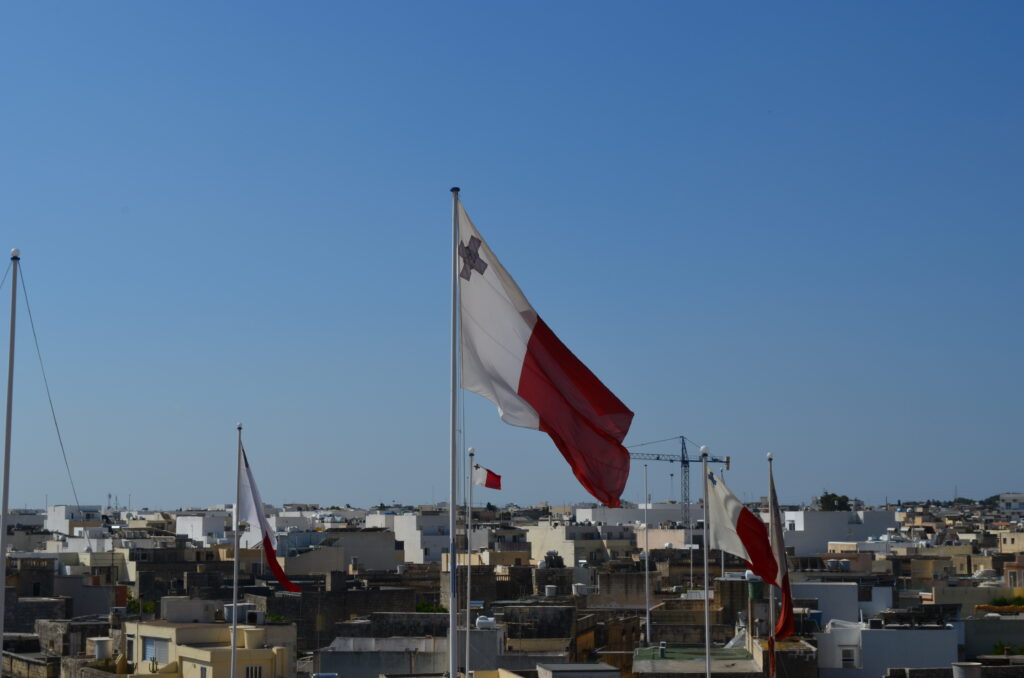 white and red flag of malta blowing in the wind. the flag has a George Cross on the upper left hand corner.