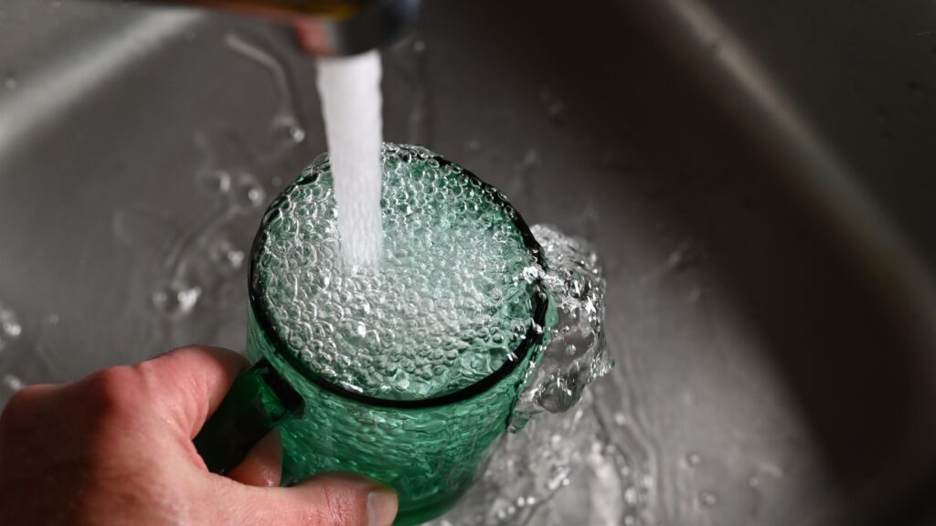 A green glass with a handle is being filled by a water tap. The water is overflowing in the glass and bubbles are forming at the top.