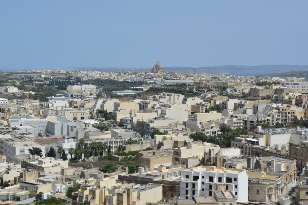 landscape photos of the island of Gozo in Malta. There are densely packed buildings in the foreground and a domed church off in the distance.