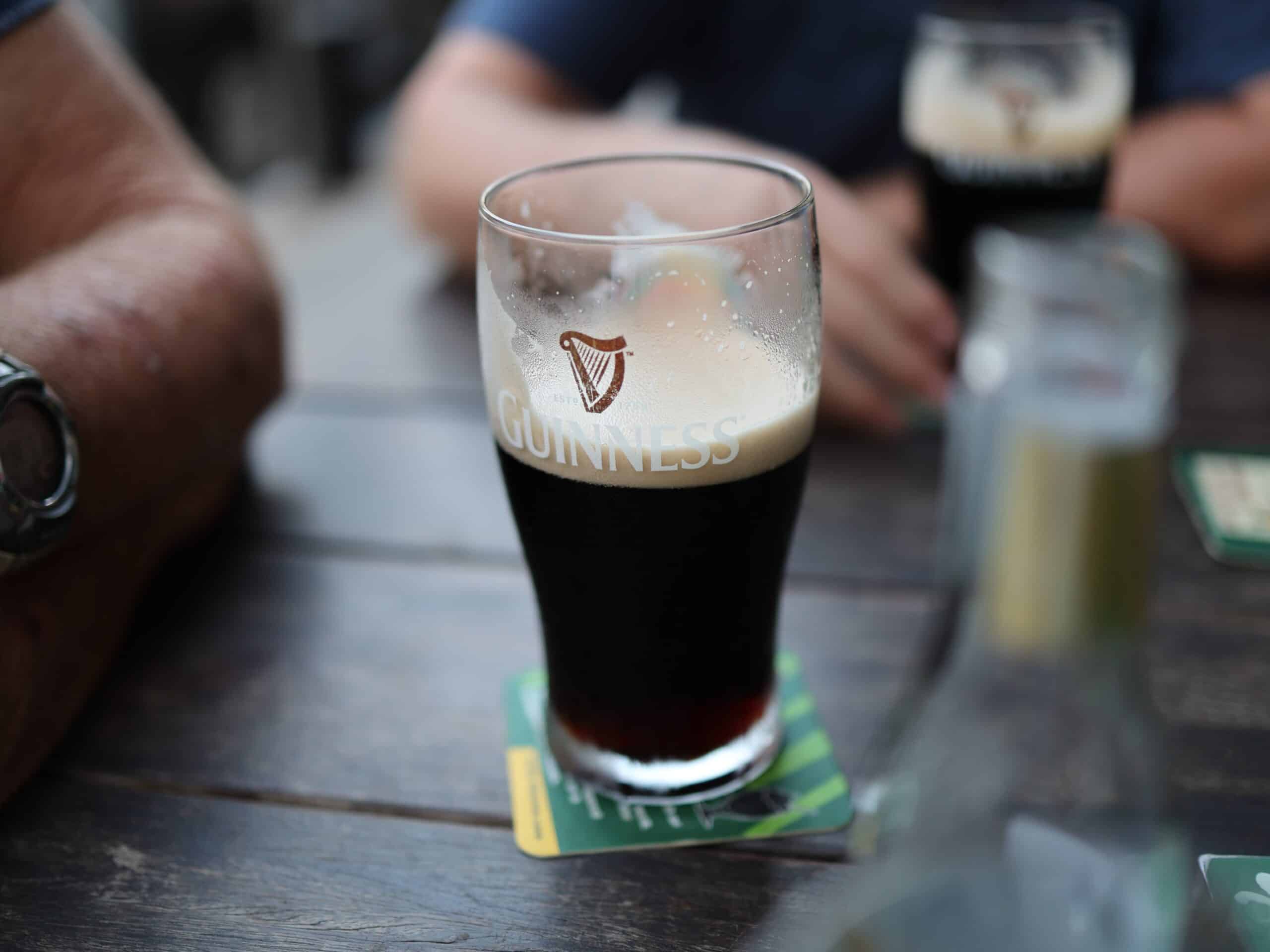 A half-full glass of Guinness beer resting on a coaster on an outdoor wooden table, with partial views of other beverages and patrons' arms in the background, suggesting a social gathering at a pub.