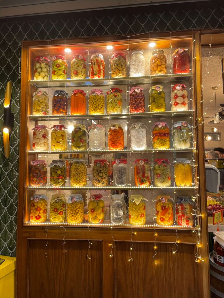 Jars of Turkish Pickles on display at a local shop. The glass jars are lined up in rows along the shelf and contain olives, corn, carrots, and other vegetables.