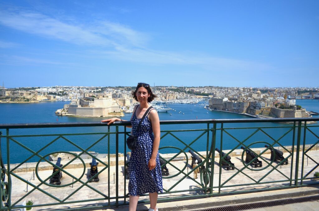 The author stands in the Upper Barrakka Gardens in the MAltese capital of Valletta. She stands in front of a black wrought iron barrier with the blue water of the harbor in the distance.