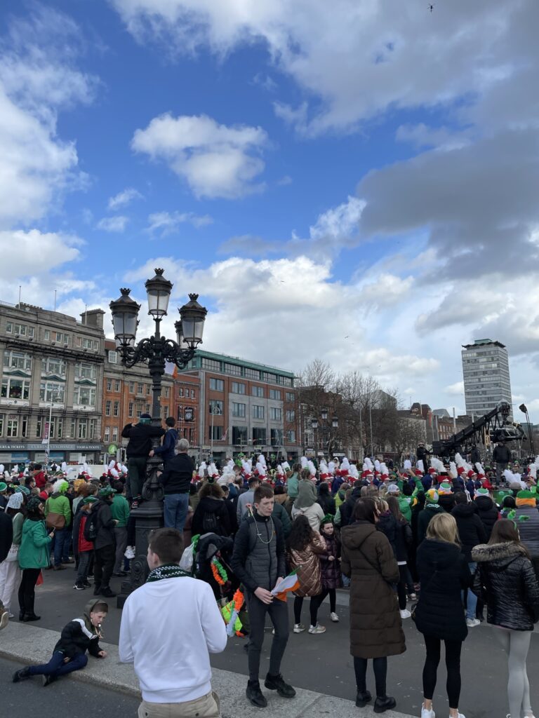 A lively gathering in Dublin, with a crowd of people, many wearing green hats, celebrating an event on a street lined with historic buildings. The atmosphere is festive, underscored by a mix of casual attire and traditional Irish accessories, against the backdrop of a partly cloudy sky.