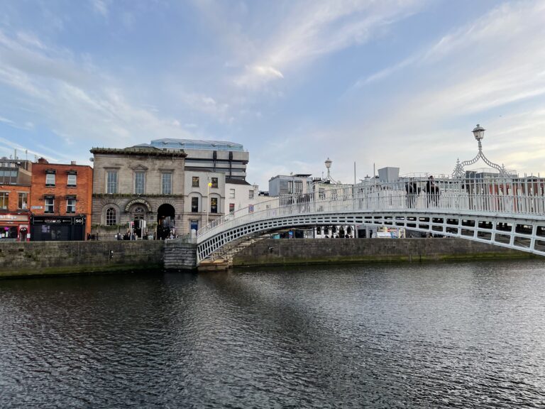 The Ha'penny Bridge arching gracefully over the River Liffey in Dublin, Ireland, on a day with partial cloud cover. The Victorian-era pedestrian bridge is captured in full view, with its white intricate railings and lampposts, connecting the lively streets lined with colorful buildings and commercial establishments.