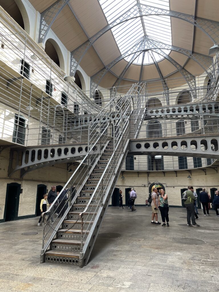 Inside the historic Kilmainham Gaol in Dublin, a striking metal staircase stands in the center, leading to the upper floors of the prison's East Wing. The Victorian architecture is evident in the vaulted ceilings and rows of arched cells, illuminated by the natural light filtering through the glass roof.