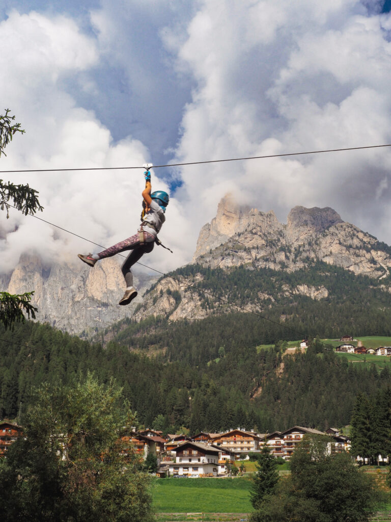 An adventurer zip-lining against a backdrop of the Dolomites' rugged peaks, with alpine houses nestled in the lush valley below, capturing the thrill of outdoor activities available in the region.