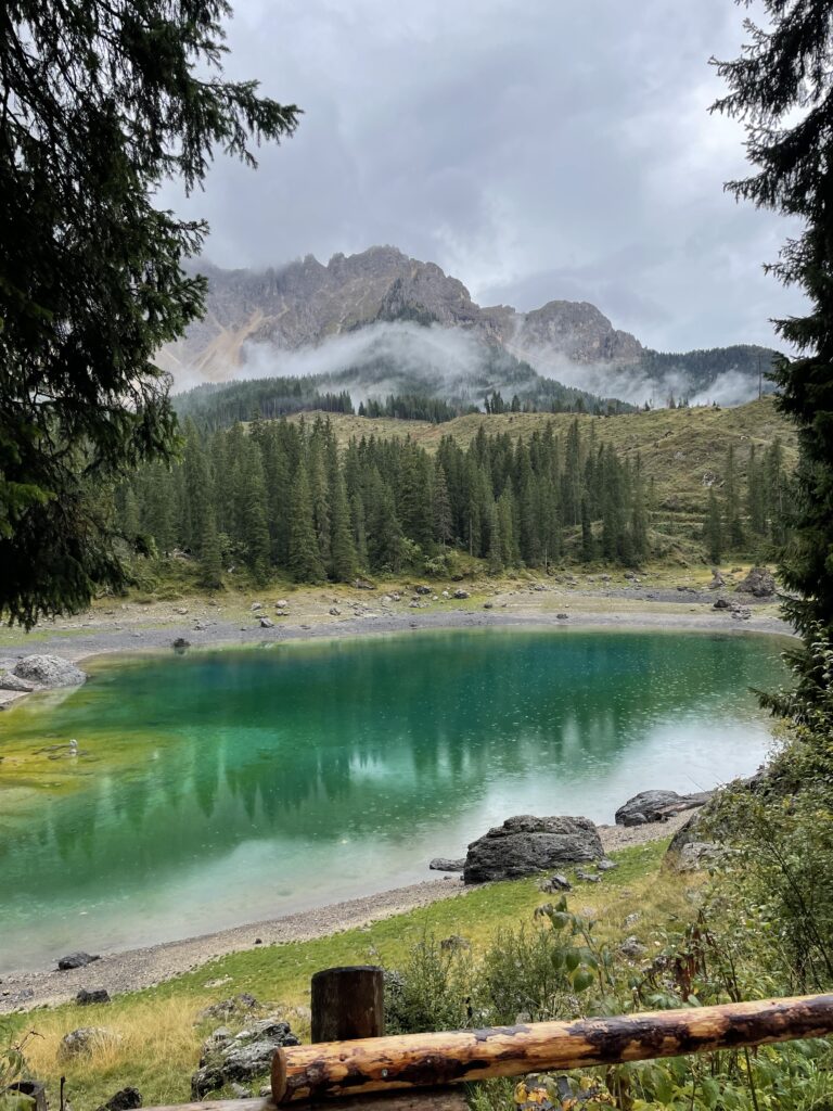 A serene view of Lake Carezza with its distinctive turquoise waters, framed by lush greenery and the misty peaks of the Dolomites, evoking a sense of tranquility and the picturesque beauty of the region.