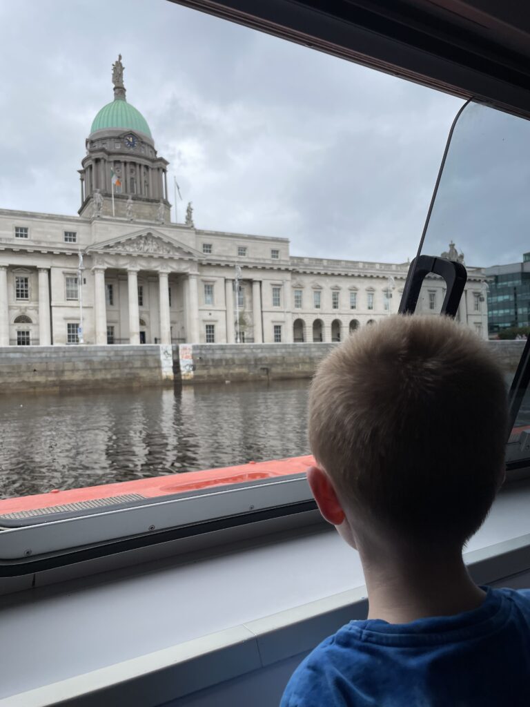 A young child with short hair gazes out of a boat window at the Custom House across the River Liffey in Dublin, with the building's prominent dome and classical architecture highlighted against an overcast sky.