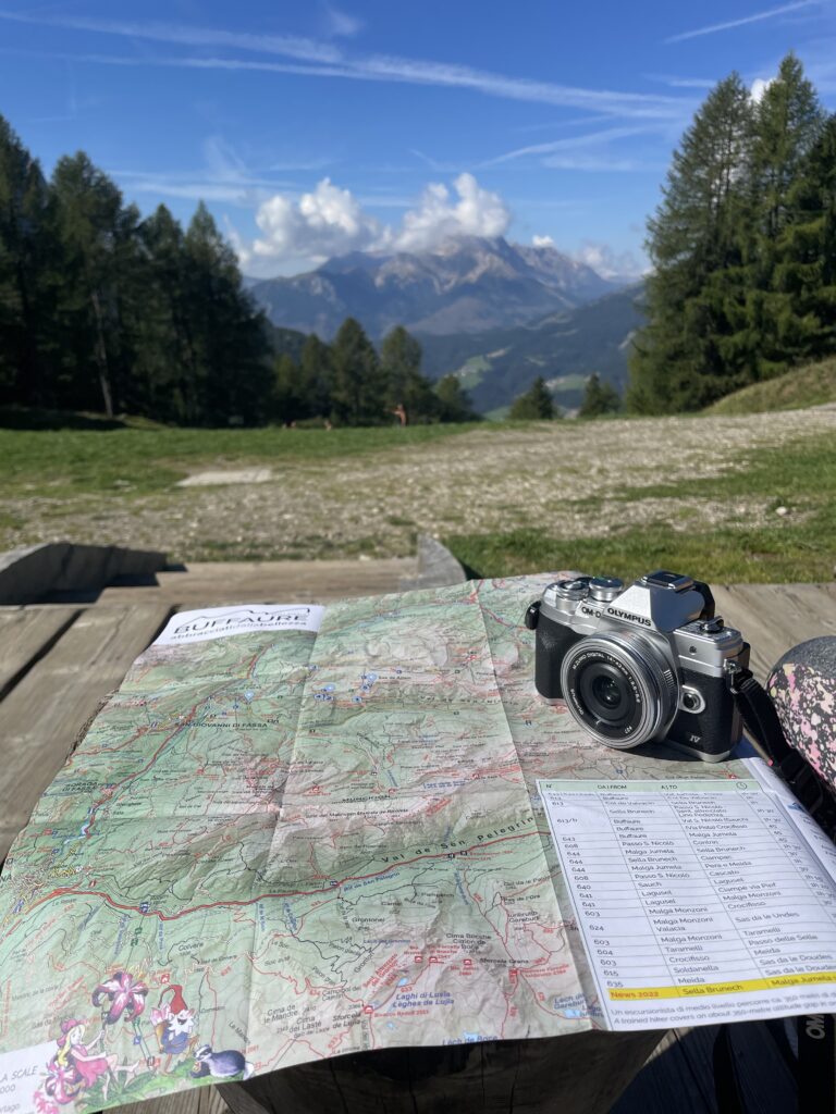 A hiking map and a camera laid on a wooden table with a blurred view of the Dolomites in the background, symbolizing the planning of an exploratory journey through the mountains.