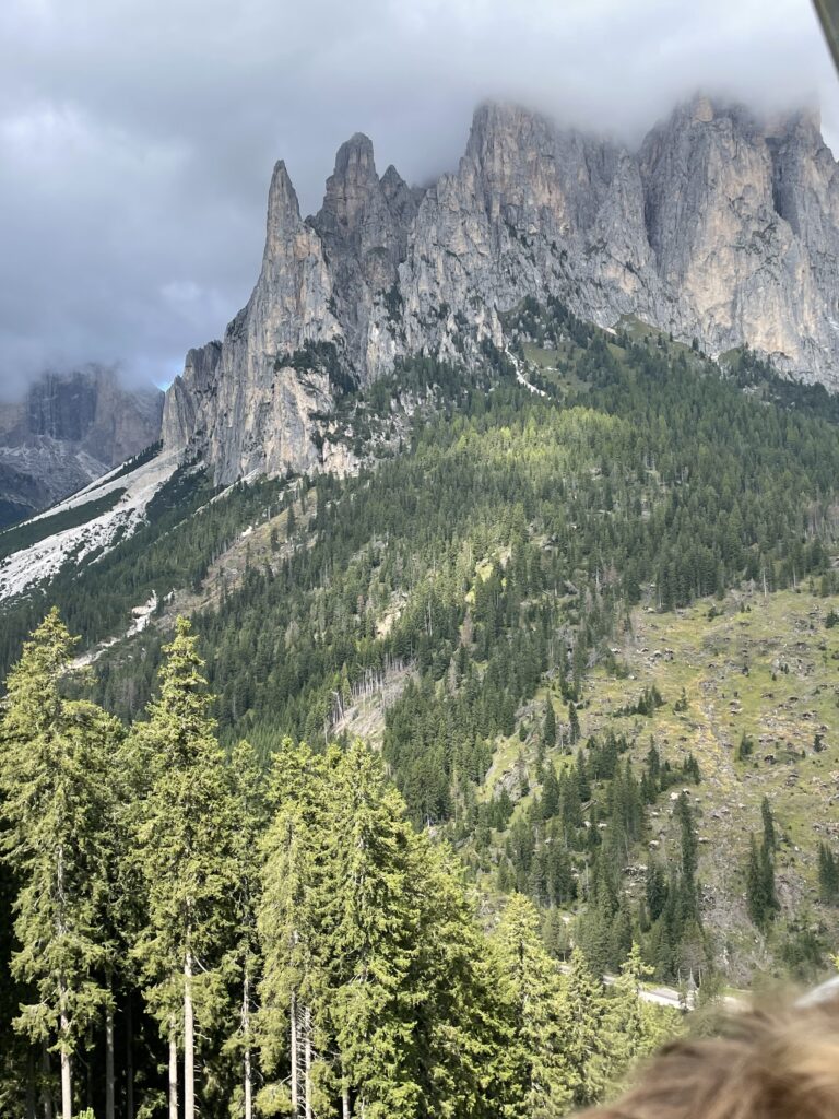 Dramatic view of the craggy spires of the Dolomites rising above a dense evergreen forest, with the peaks partially enshrouded by low-hanging clouds, showcasing the region's striking alpine landscape.