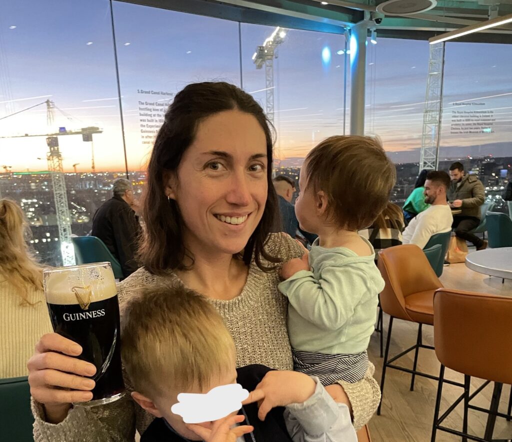 The author, Tamar, smiling at the camera, holding a pint of Guinness in a glass with the brand's logo, while in the background is the panoramic view of Dublin's skyline at dusk from the Gravity Bar at the Guinness Storehouse. She is holding two children, with one's face obscured for privacy, indicative of a family moment enjoying the city's famous landmark.