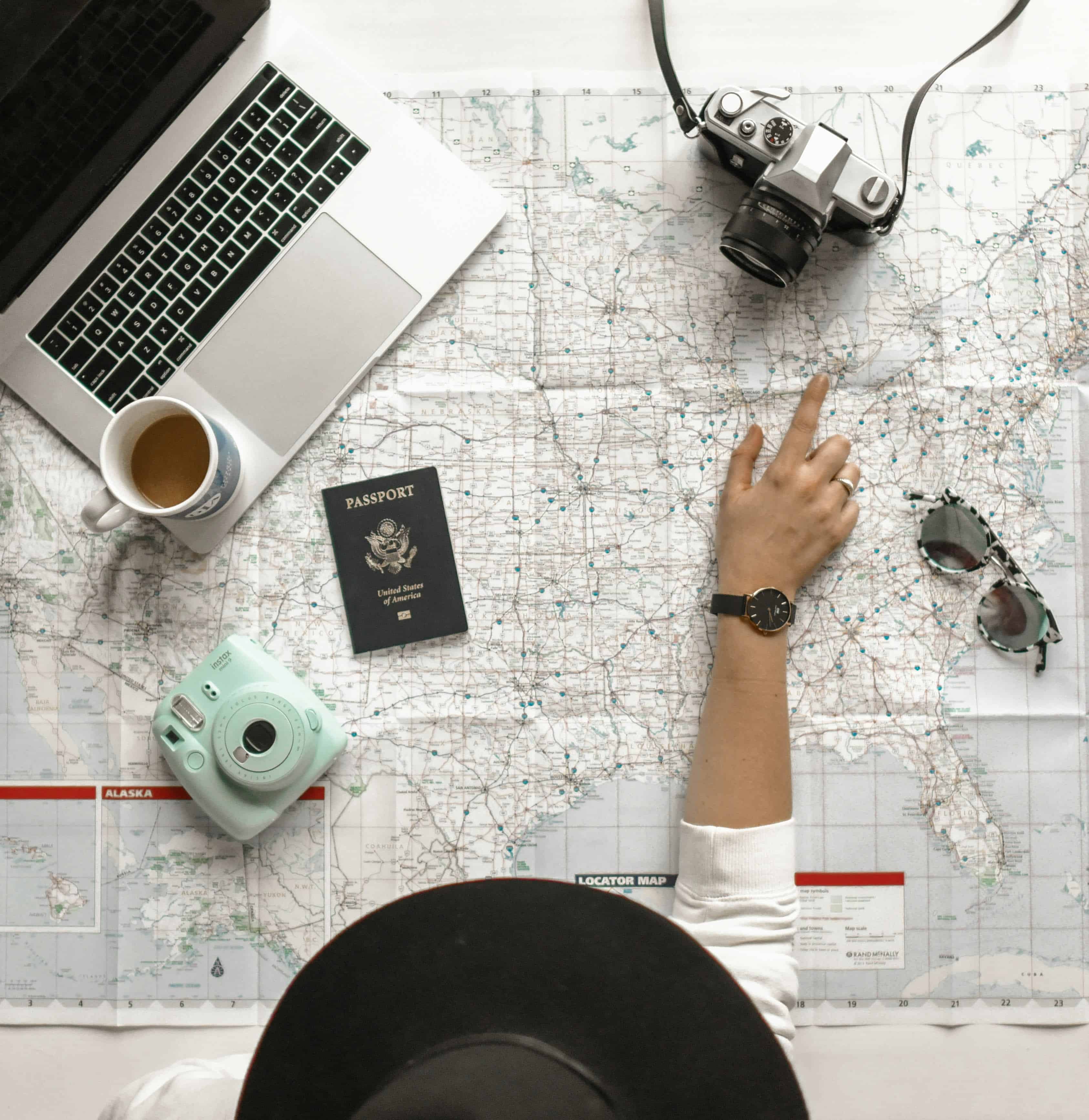 Overhead view of a weekend getaway planning scene with a map of the United States, a laptop, camera, sunglasses, and a passport, with a person's hand pointing to a location on the map.