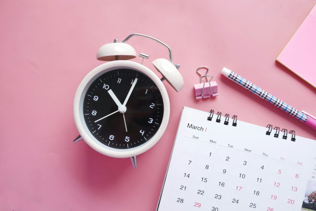 A classic white alarm clock showing ten past ten, with a pink binder clip and a pen, next to a calendar open to March with days marked, all on a pastel pink background.