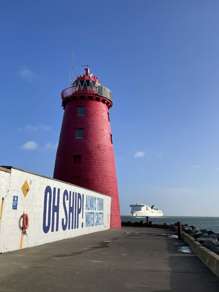 Poolbeg Lighthouse painted in vivid red, standing at the end of a long pier against a clear blue sky. To the left, a white wall features the humorous safety message 'OH SHIP ALWAYS THINK WATER SAFETY' in bold blue lettering, with a ship visible on the horizon, emphasizing the nautical theme of the scene.