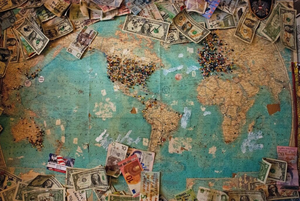 A weathered world map pinned with various currencies and colorful thumbtacks, indicating different locations and travels, symbolizing global finance and tourism.