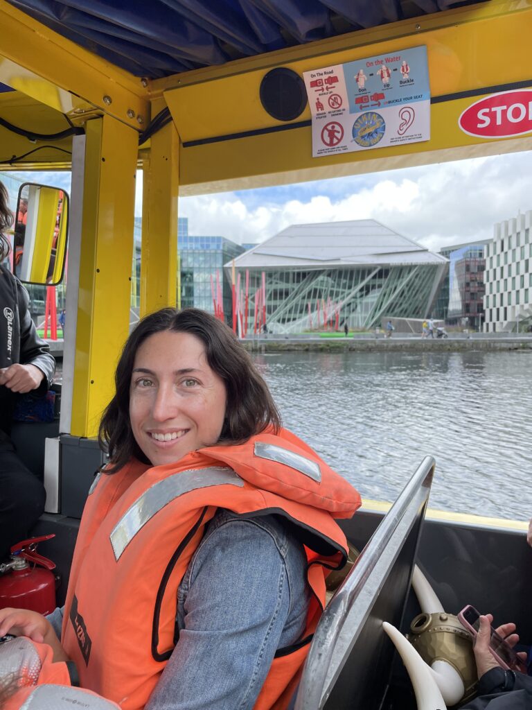 The author, Tamar, wearing an orange life vest smiles while seated on a Viking Splash Tour amphibious vehicle, with Dublin's modern glass-fronted buildings and calm water in the background.