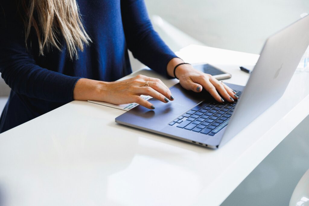 A person booking their weekend trip accommodations on a laptop with their left hand on the trackpad and right hand on a smartphone, both placed on a white desk, showcasing a multitasking work environment.