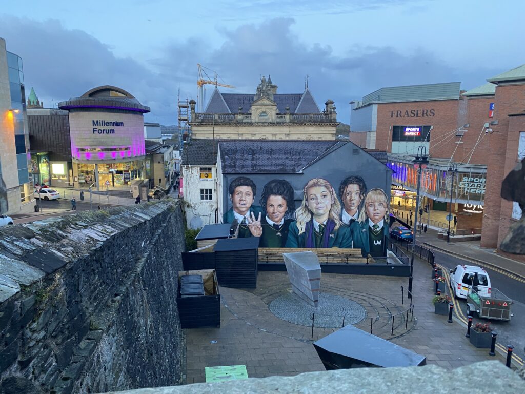 Twilight scene in Derry featuring a mural of the 'Derry Girls' TV show characters on a building wall, with the Millennium Forum and surrounding streets lit up in the background.