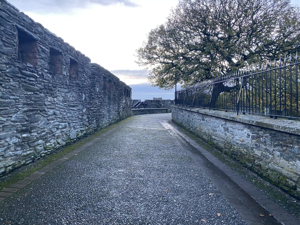 A historical perspective of the Derry Walls in Northern Ireland, showcasing the rugged stone texture and the ancient defensive structure, bordered by a metal fence and bare trees against a dusky sky.