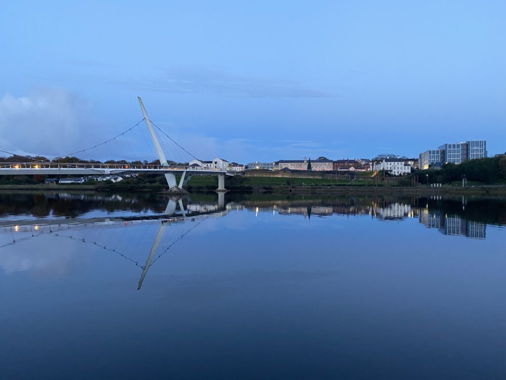 Calm evening view of the River Foyle in Derry, Northern Ireland, with the Peace Bridge reflecting on the water and the cityscape gently illuminated at twilight.