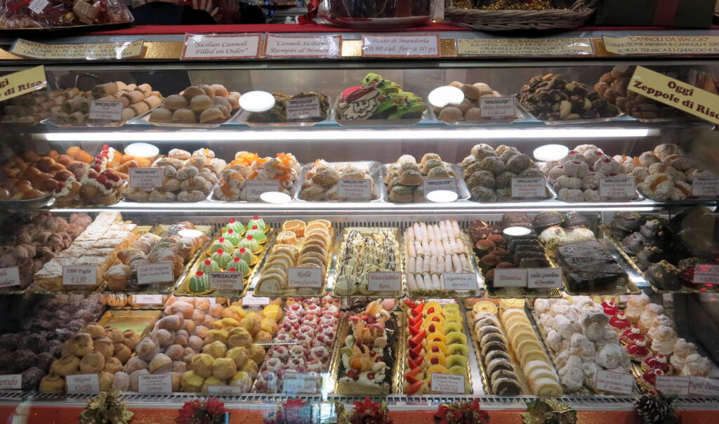 A vibrant display of various Sicilian pastries in a bakery case, including Cannoli, almond pastries, and colorful fruit-topped desserts, with price tags and descriptions in Italian.