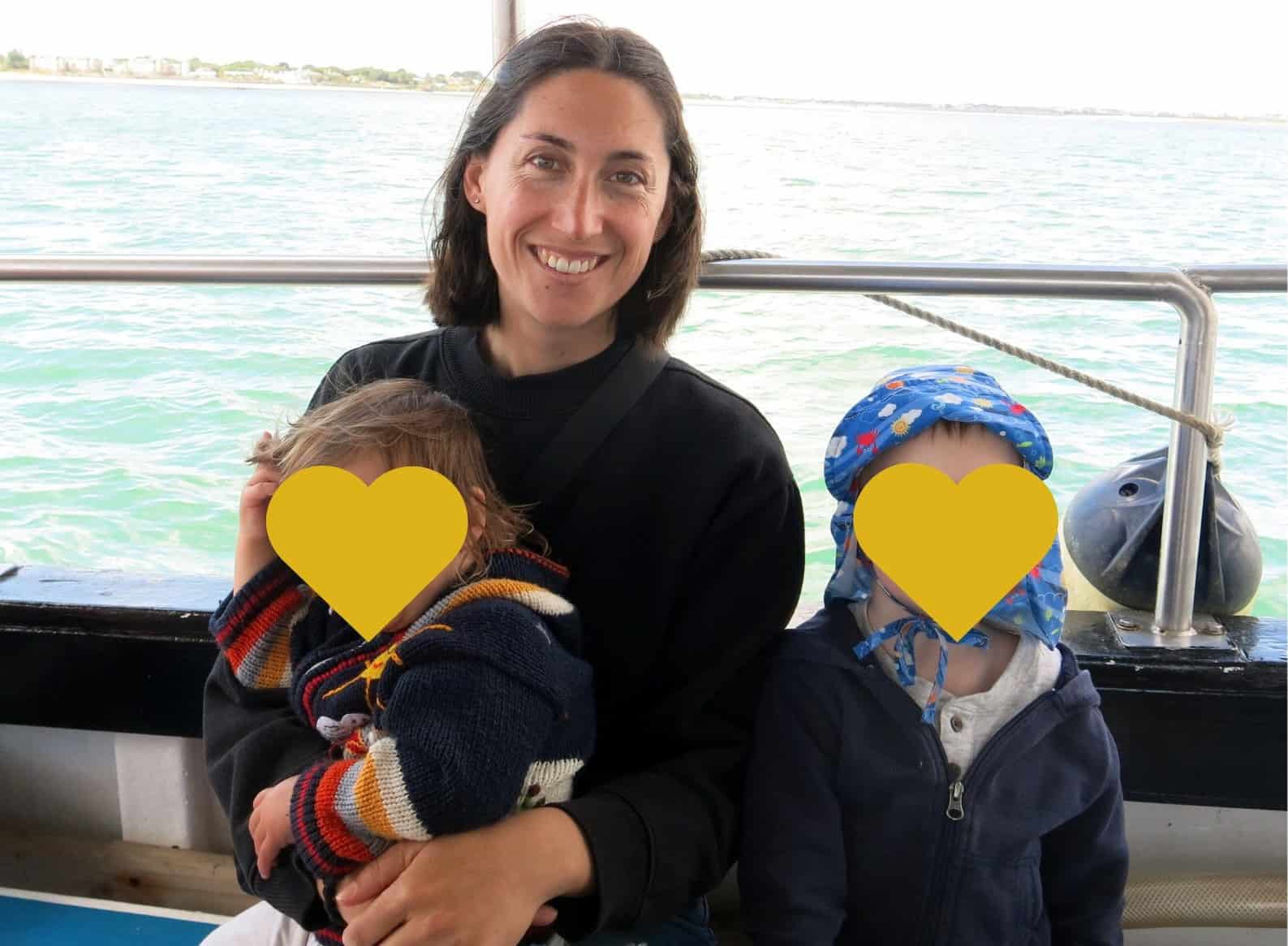 A smiling woman, the author, with dark hair, wearing a black sweater, is on board the Ireland's Eye Ferry, embracing her two young children whose faces are covered by heart emojis for privacy. They're near the ferry's railing with a glimpse of the calm sea and the coastline in the distance, capturing a family moment on a coastal excursion.