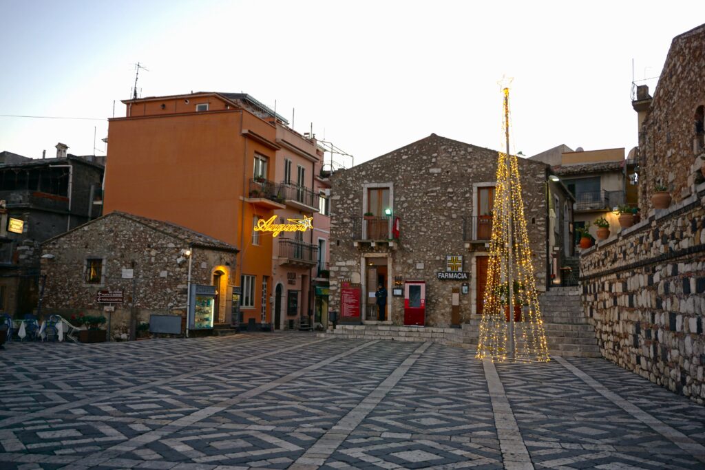 A quaint square in Castelmola, Italy, during dusk with a modern Christmas tree made of lights, stone buildings with a mix of vibrant and rustic facades, and a clear sky transitioning from day to night.