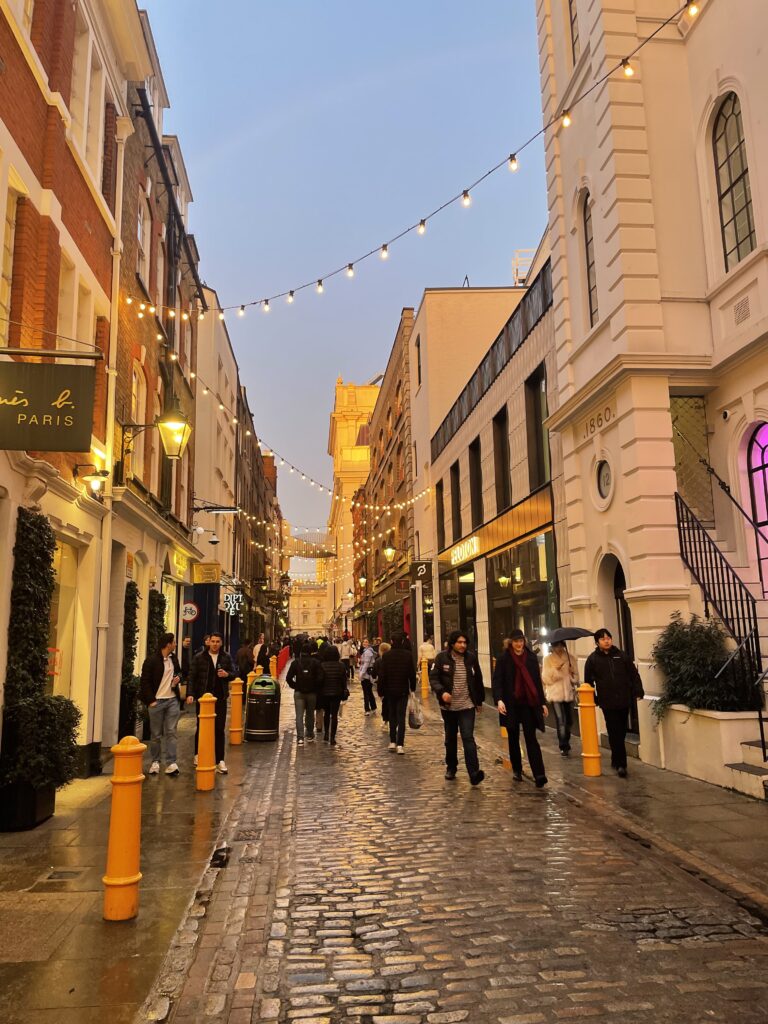 Pedestrians stroll through a cozy London street at dusk, with twinkling Christmas lights strung above, wet cobblestones reflecting the warm glow, and historic buildings lining the path.