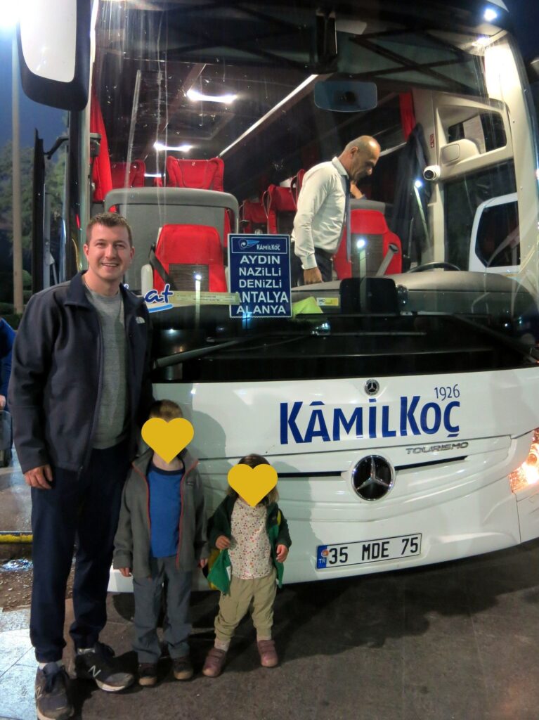 A smiling man and two children, whose faces are obscured for privacy, stand in front of a Kamil Koç bus at night. The destination board on the bus lists Turkish cities like Aydin, Nazilli, Denizli, Antalya, and Alanya.